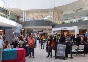 Job fair taking place in a shopping mall, with tables staffed by employers, attendees visiting booths, and signs identifying different organizations.