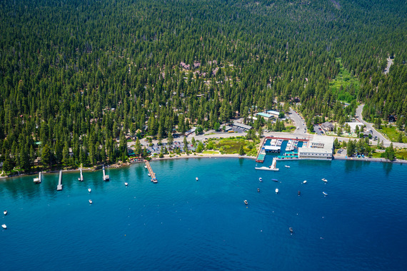 An Aerial photo of Carnelian Bay, multiple deck boats, and docks with the road, houses, businesses, and green forest nearby
