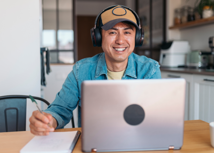 A man working on a laptop and writing on a notepad, smiling toward the viewer