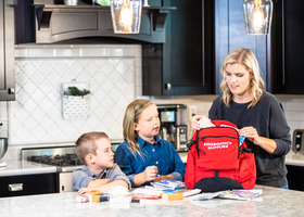 Mom and two kids in a kitchen packing a red backpack with emergency supplies to prep a go-kit.