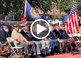With a play button overlay, several uniformed honor guard members stand on a track holding the flags of America’s armed services.
