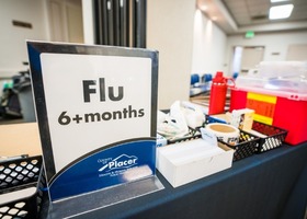 Sign indicating flu vaccinations available for individuals aged six months and older at a health clinic table setup.