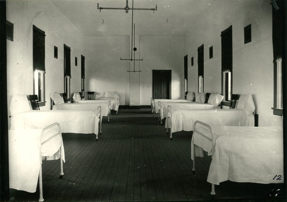 Two rows of neat hospital beds line the walls of a building hall with windows