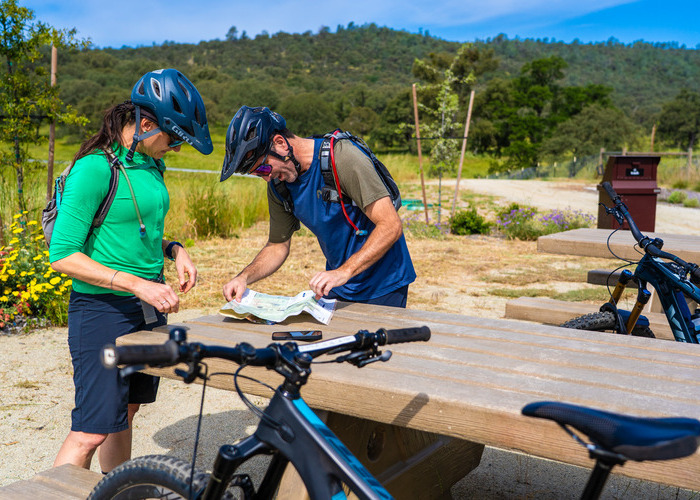 Two bikers rest and review a map with bikes against a picnic table near Hidden Falls Regional Park and green grass.