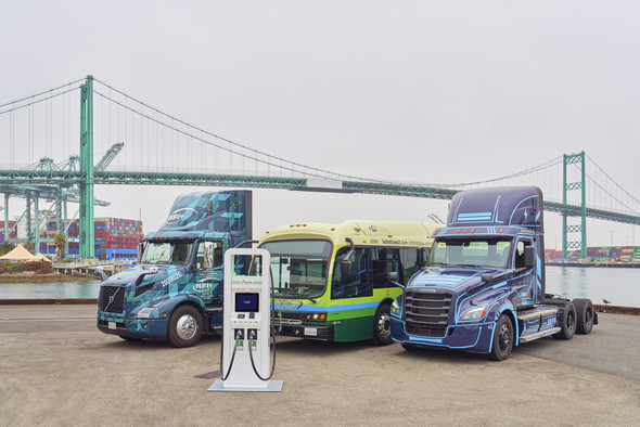 Two zero-emission trucks and zero-emission parked behind a charging station and in front of a port backdrop.