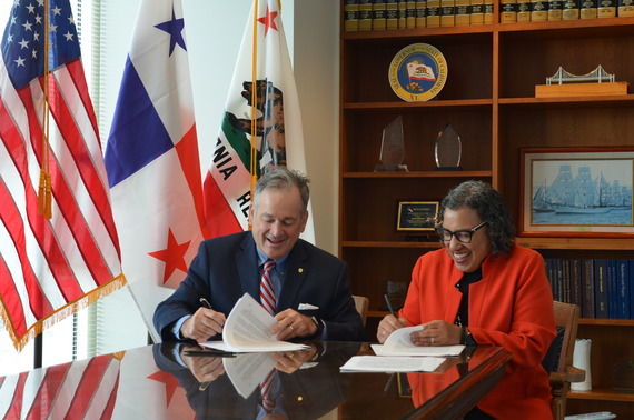 Two officials, seated at a conference table, smile as they sign a formal agreement. 