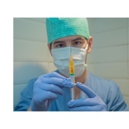 A medical worker wearing scrubs, gloves, a mask and a surgical cap holds a syringe filled with orange liquid. 