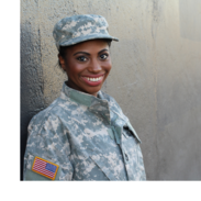 A woman wearing military fatigues stands against a wall and smiles.