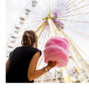 A woman holds pink cotton candy and looks up at a Ferris wheel.