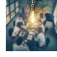A closeup of a lit lightbulb. In the background, a conference table full of business people deep in conversation.
