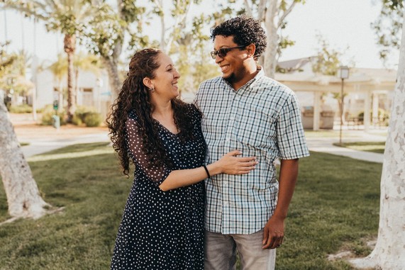 A woman smiles and hugs a man. They both stand outside in a park in the daytime. 