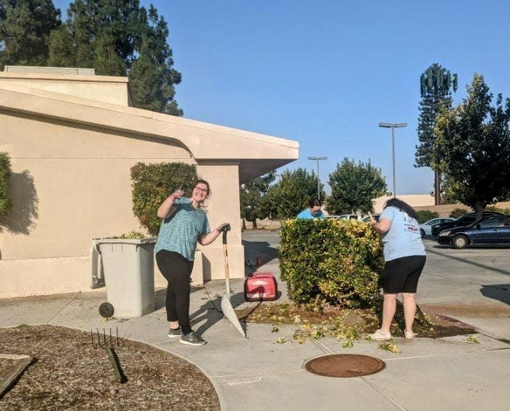 Two girls stand in front of a bush that has been trimmed. They are shoveling the trimmings. One girl turns to the camera and gives a thumbs up.