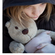 A young girl sitting on the ground outside and wearing a hoodie clutches a teddy bear tightly to herself.