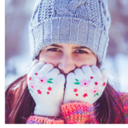 A girl standing outside in the snow wears a knit hat and holds her hands to her face. She is wearing mittens.