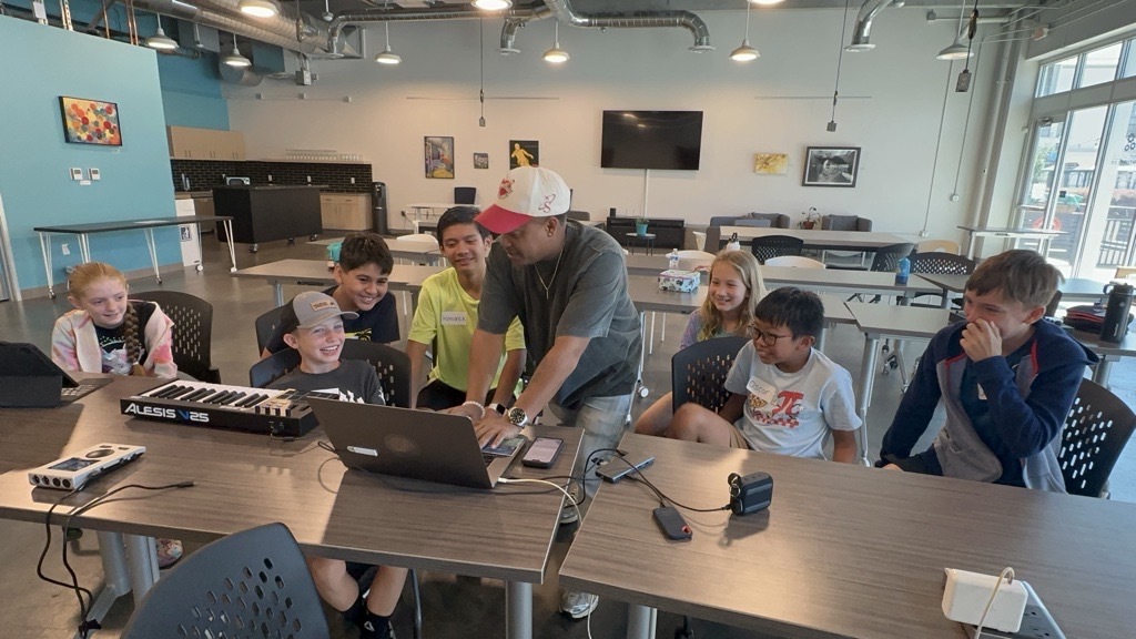 Photo of an instructor teaching 7 smiling children how to make music on a laptop