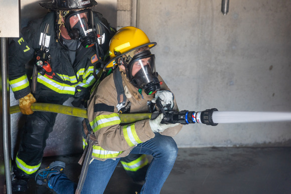 Photo of a man and a child, both dressed in fire department protective gear. Both are looking in the distance as they operate an active fire hose.