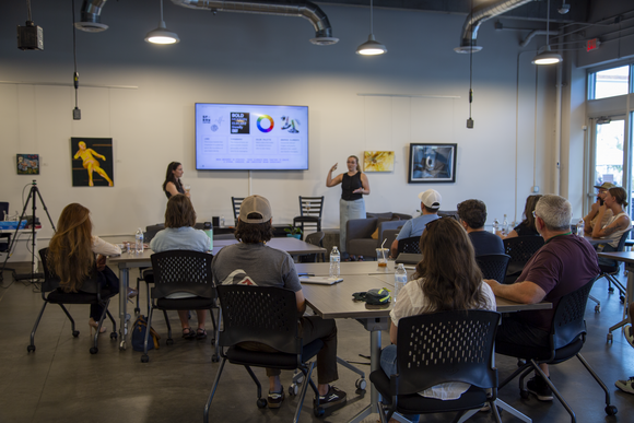 Two individuals stand in front of a class, giving a presentation while pointing to the slideshow on a wall-mounted TV screen.