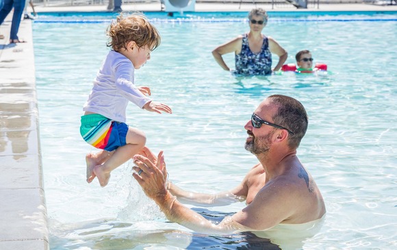 A toddler jumps into a pool, where his father smiles with his arms extended to catch him