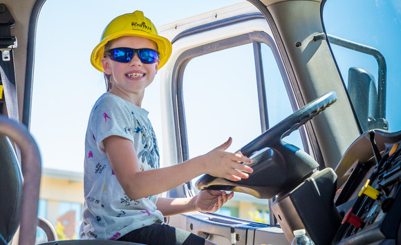 A girl wearing a construction hat and sunglasses smiles while sitting in the driver seat of a utility truck