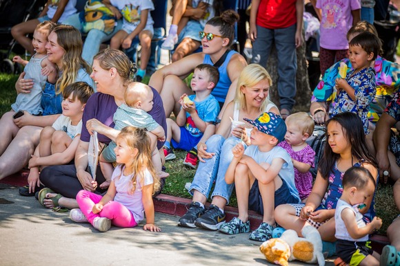 A crowd of parents and their children smile and chat as they sit on a sidewalk