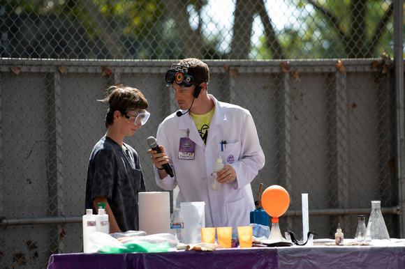 A teacher in a lab coat holds a microphone up to a student as they stand behind a table with a lab demonstration