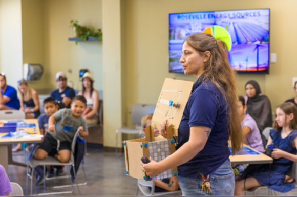 A teacher presents a model of a building with a pie chart showing Roseville energy sources on a screen behind her