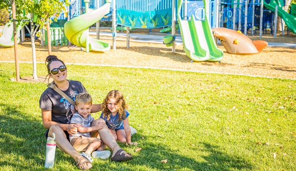 A woman smiles and sits with two children on her lap in Weber Park. A playground can be seen in the background.