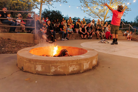 A back-side view of a presenter raiseing their arms as they speak to a crowd. An active campfire is in the foreground.