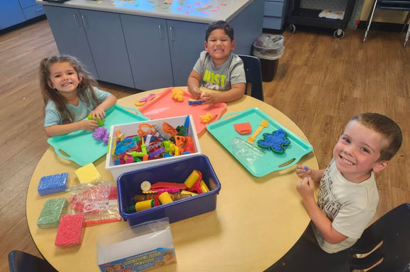 Three preschool students smile while playing at a round table