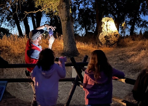 Two children watch a Rock Art Mysteries tour guide shine a light on a boulder