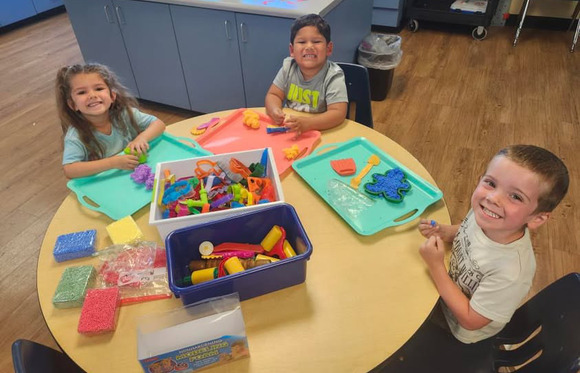 Three preschoolers smile while completing a craft at a table