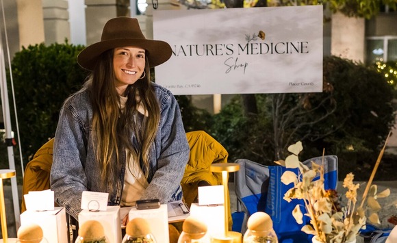 A woman stands at the booth of her small business, "Nature's Medicine" as she smiles with her inventory