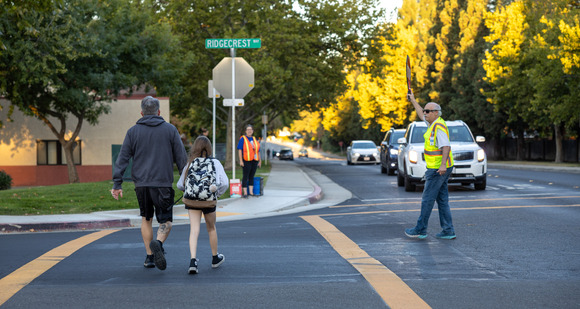 An adult walks with a child on a cross walk as a crossing guard directs traffic.