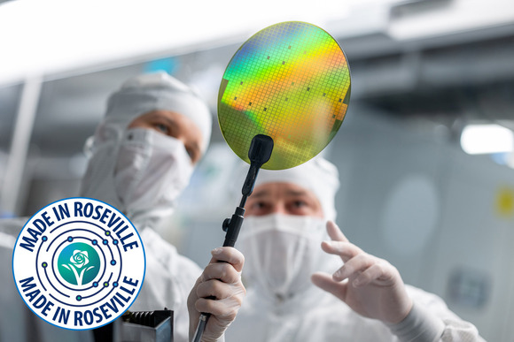 Two people in lab suits and masks hold up a semiconductor chip with forceps. The Made In Roseville logo in in the corner.