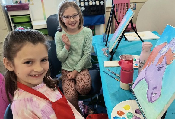 Two children smile next to their art stations and portraits.