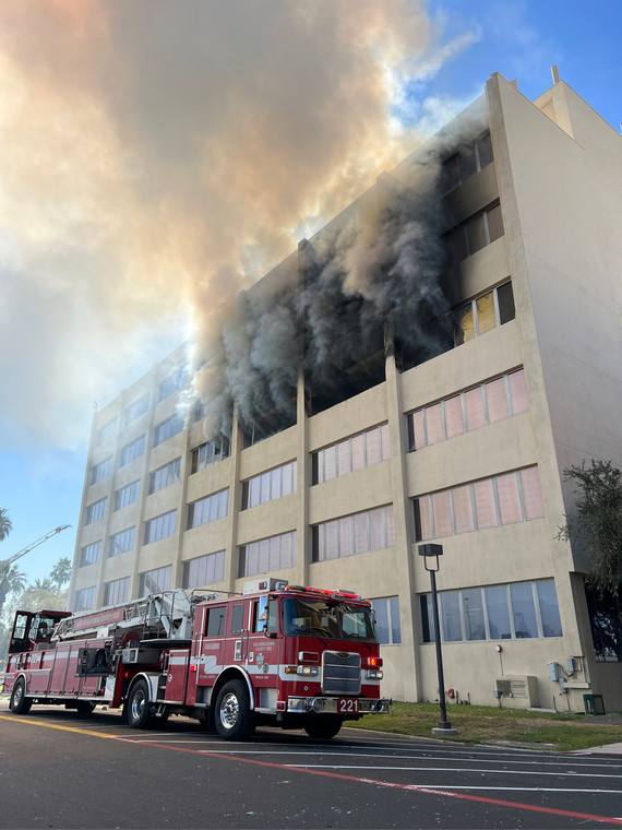 A picture of a six-story burning building with black smoke coming out of the top three floor windows. A fire truck is in front.
