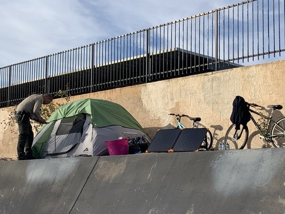 A sheriff deputy is seen leaning into a homeless perspmtent on the side of a wash with trash and a bicycle.