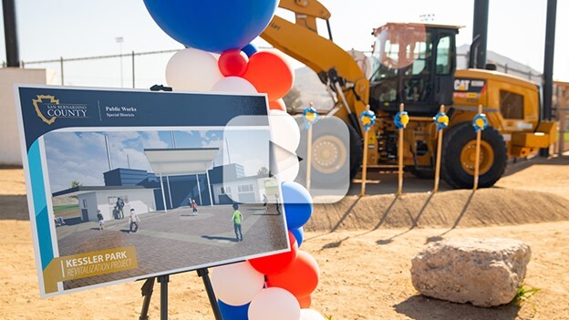 A photo of a dirt ground with construction equipment behind it and a easel in the foreground with a project rendering on a new building.