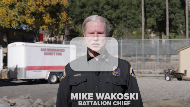 A male fire battalion chief is seen speaking to the camera with a fire trailer and country landscape in the background.