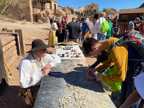 A group of kids stand at a table learning how to shape and sharpen an arrowhead out of rock on a brick at Calico California Days.
