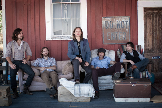 A group of male musicians posing in front of a red barn type building sitting on an old couch and and two old recliners.
