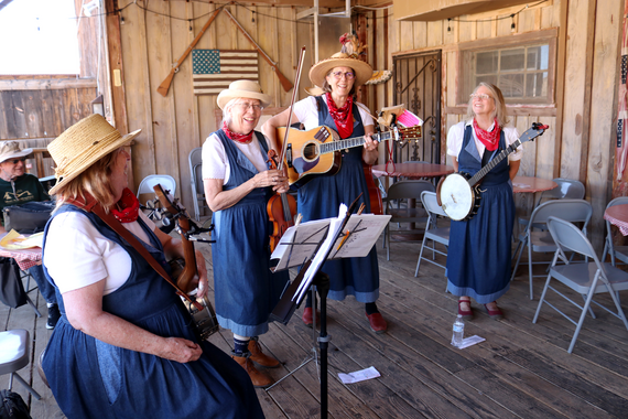 An all-female group of musicians holding a guitar, banjos and fiddles in blue denim dresses on the porch of Calico Restaurant.