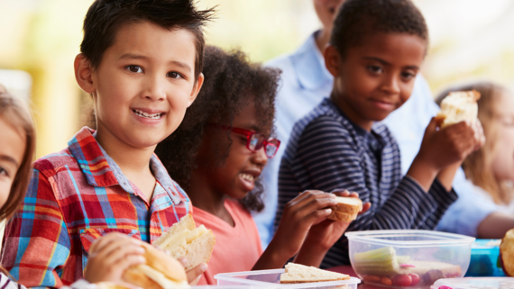 A photo of a young boy smiling while sitting at a table with two other children: a bot and girl eating lunch.