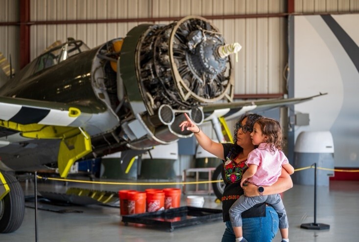 A woman holding a young girl in her arms point in the air with a vintage plane behind her in a airport hangar.