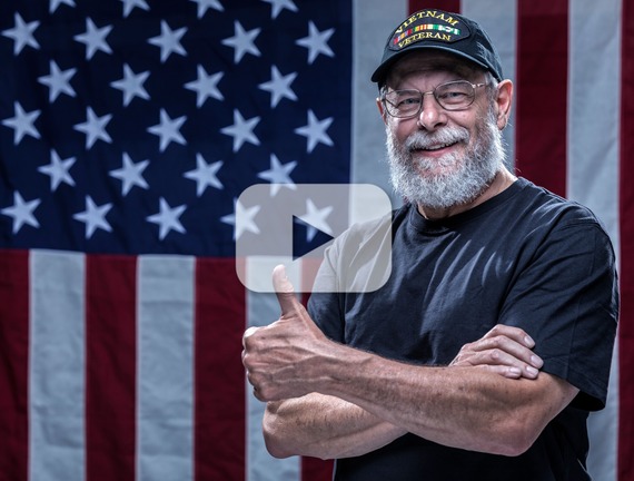 Person showing a thumbs up in front of an American flag, wearing glasses and a black cap that says Vietnam Veteran.