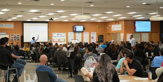 A large group of people sitting in a meeting room listening to a speaker presenting at the front near a projector screen. 