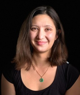 Portrait of a smiling person wearing a black top and a green pendant necklace set against a dark background.