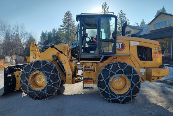 A yellow Caterpillar 930-wheel loader equipped with heavy-duty snow chains on its tires is parked outdoors. 