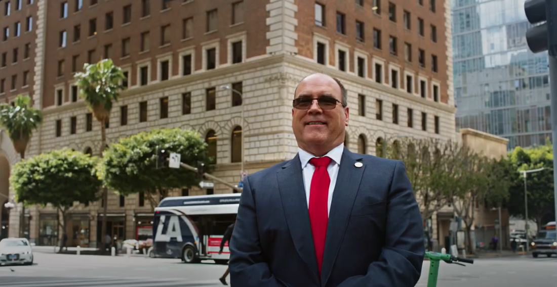Man in suit and red tie stands on city street corner with LA bus, palm trees, and historic brick building in the background.