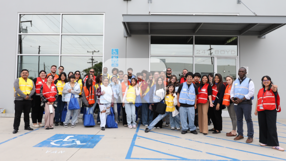 A diverse group of smiling people stands outside a building wearing colorful vests. Some hold blue tote bags.  